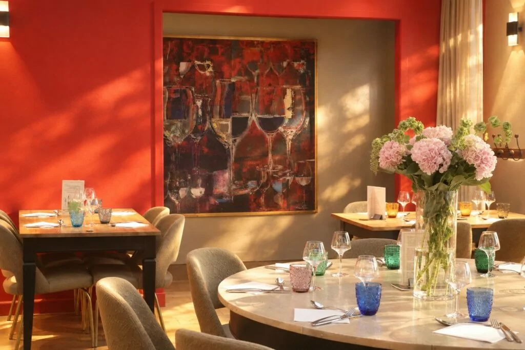 Salle du restaurant : mur rouge, grand tableau de verres de vin, table ronde dressée et bouquet d'hortensias dans la lumière du soir