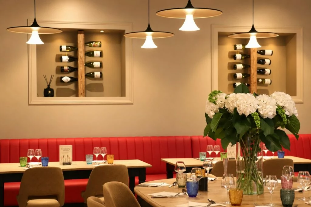Salle du restaurant : longue banquette rouge, niches de bouteilles au mur, suspensions et bouquet d'hortensias blancs sur une table ronde